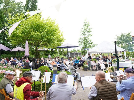 Musiker einer Blaskapelle spielen im Vordergrund im Freien. Dahinter sitzt ein großes Publikum auf Stühlen in einem Garten oder Park und genießt das Konzert unter Bäumen und Zelten.