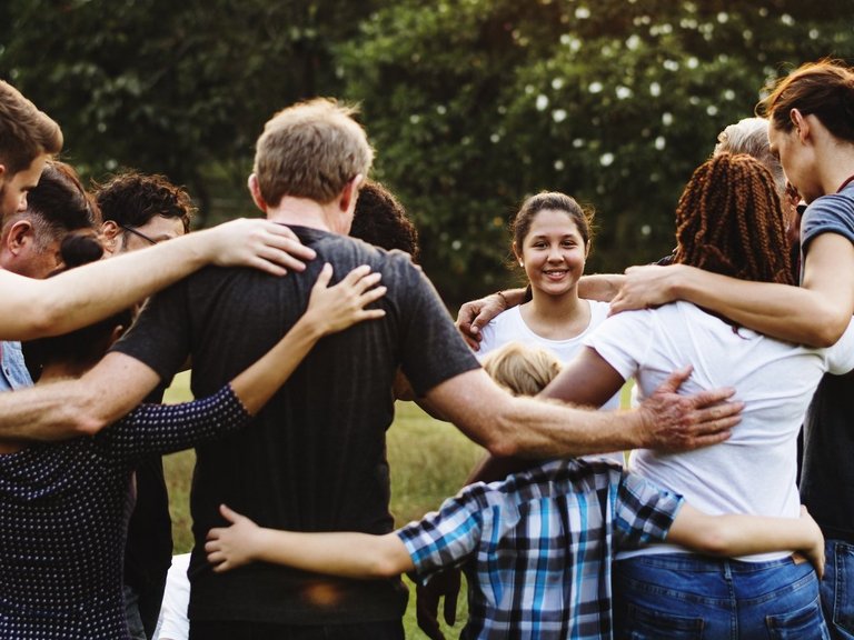 Mehrere junge Menschen stehen im Kreis und fassen sich an den Schultern, die Köpfe gesenkt zum Kreisinneren gewandt. Ein Mädchen blickt zum Betrachter und lächelt.