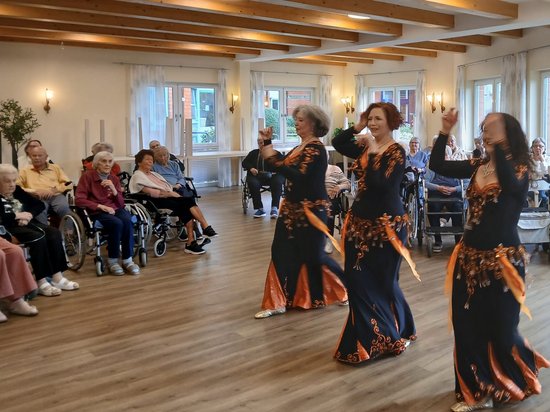 Drei Frauen in dunklen, orangefarben gemusterten Kleidern tanzen vor einem Publikum aus älteren Menschen. Viele Zuschauer sitzen in Rollstühlen in einem hellen Saal mit Holzbalkendecke und beobachten die Aufführung.